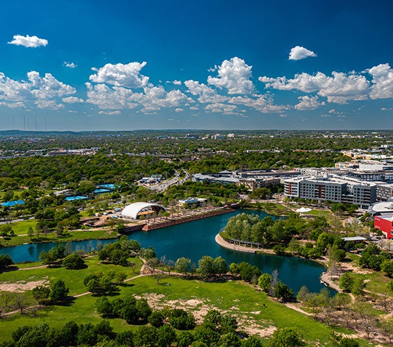 High-angle view of Mueller Lake Park in Austin, Texas, showcasing green open spaces, walking trails, a central lake, the outdoor amphitheater, and surrounding modern residential and commercial buildings under a vibrant blue sky with scattered clouds.