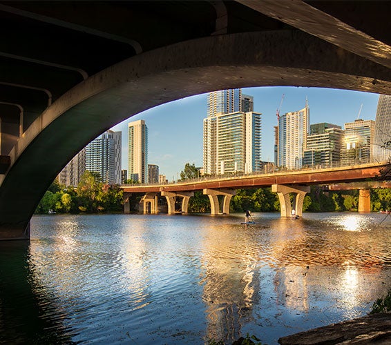 Dramatic view of downtown Austin, Texas, seen through the arches of a bridge at sunset. The skyline is reflected in the calm waters of Lady Bird Lake, with paddleboarders enjoying the scene beneath the South First Street bridge.