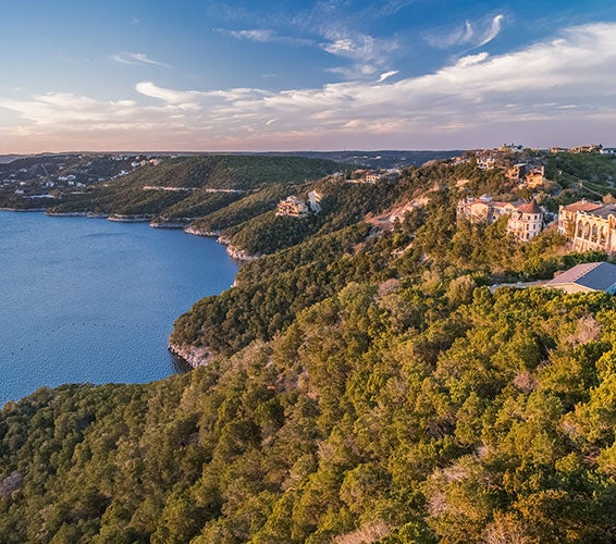Aerial view of luxury homes perched on the hillside above Lake Travis in Austin, Texas, with sweeping views of the lake and Hill Country landscape under a vibrant sunset sky—highlighting premier waterfront real estate in West Austin.
