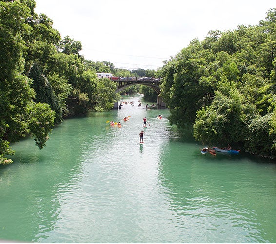 Scenic view of people paddleboarding and kayaking along the clear green waters of Barton Creek in Austin, Texas, surrounded by dense trees and greenery with a historic arched bridge and traffic visible in the background.