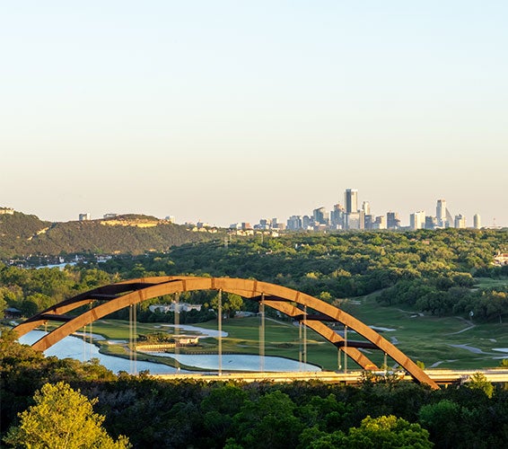 Sweeping view of the Pennybacker Bridge (360 Bridge) spanning Lake Austin, with a backdrop of rolling Hill Country greenery and the distant downtown Austin skyline glowing in the early evening light.