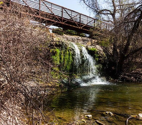 Small scenic waterfall cascading beneath a wooden pedestrian bridge along the Brushy Creek Trail in Cedar Park, Texas—highlighting the natural beauty, hiking, and outdoor recreation opportunities in North Austin suburbs.