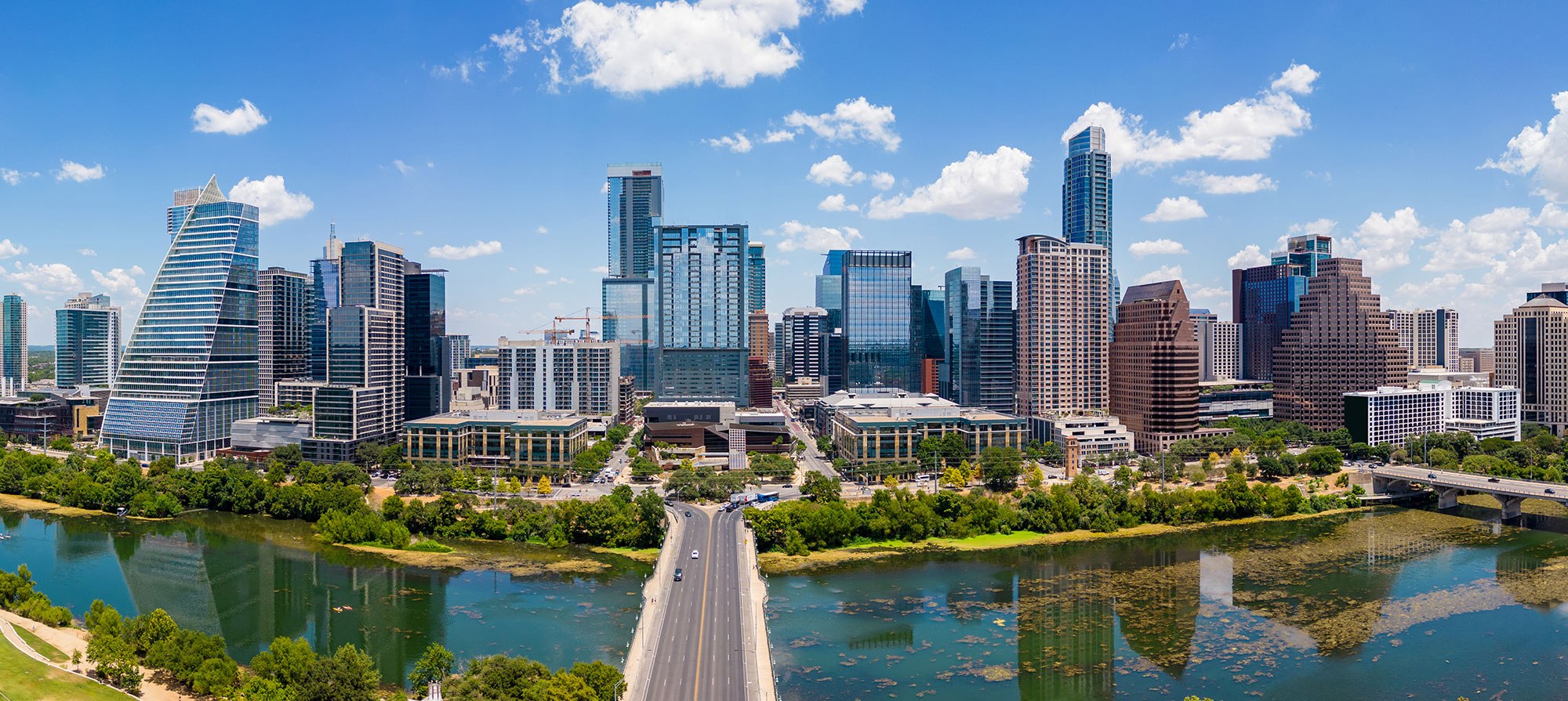 Aerial skyline view of Downtown Austin, Texas, showing the Colorado River, Ann W. Richards Congress Avenue Bridge, and modern high-rise condos in the 78701 area under a bright blue sky.