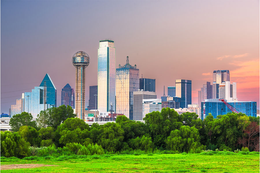 Vibrant view of the Dallas, Texas skyline at sunset, featuring the iconic Reunion Tower, Bank of America Plaza, and other reflective skyscrapers rising above a lush green foreground with trees and open field.