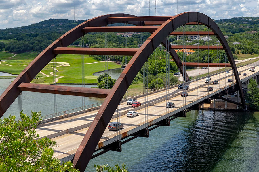 Aerial view of the Pennybacker Bridge (360 Bridge) in Austin, Texas, spanning across the Colorado River with cars driving across and lush green hills and a golf course in the background.